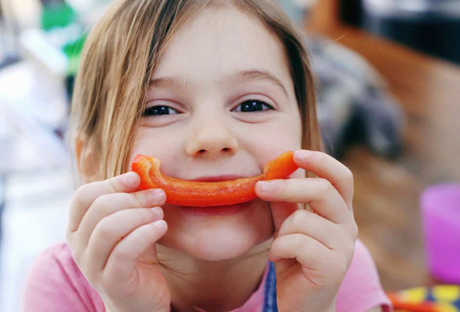 A young girl smiles while holding up a piece of red pepper up to her mouth. The piece of pepper recreates a smile.