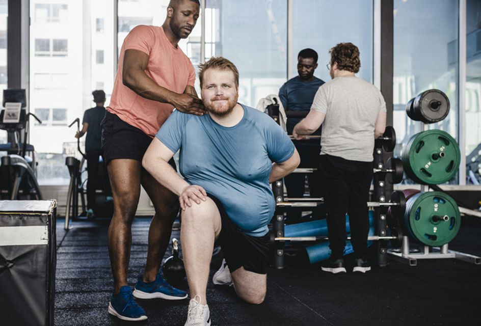 A coach in a gym helps a man position his body correctly while doing a lunge.