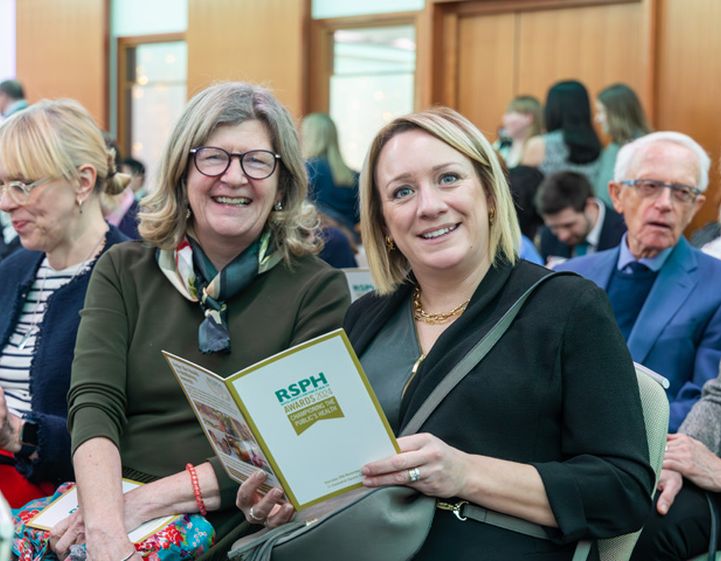 Two women sitting down at a busy Health & Wellbeing Awards ceremony smiling to camera with one holding an event programme.