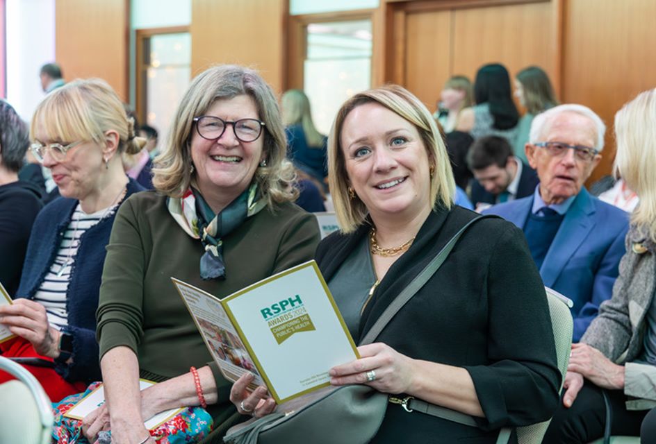 Two women sitting down at a busy Health & Wellbeing Awards ceremony smiling to camera with one holding an event programme.